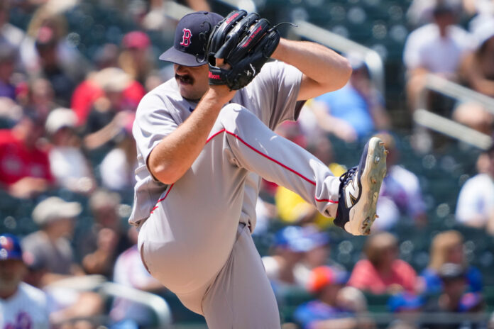 Baseball pitcher in gray uniform delivering a pitch with his glove up near his face, crowds blurred in the background.