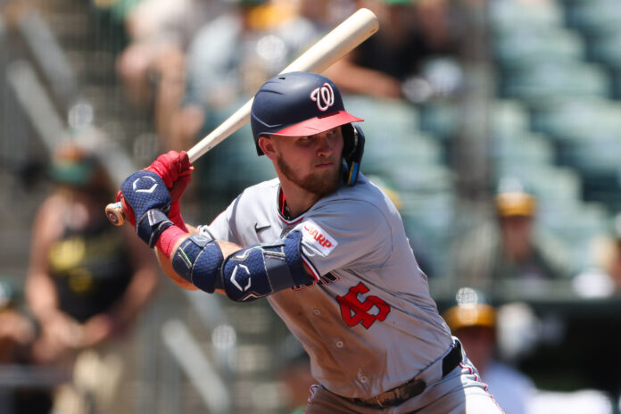 Baseball batter in a Nationals uniform swinging a bat during a game, with a helmet and red gloves.