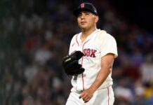Suarez Return Delayed: Red Sox Must Trust the Process Now Baseball player in a Red Sox white uniform with a black glove on the field, looking ahead during a game.