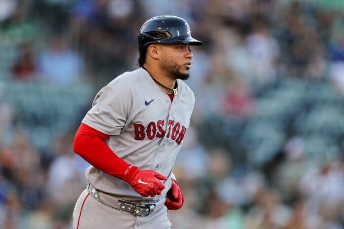 Baseball player in a Boston uniform running on the field with a helmet and red sleeves.