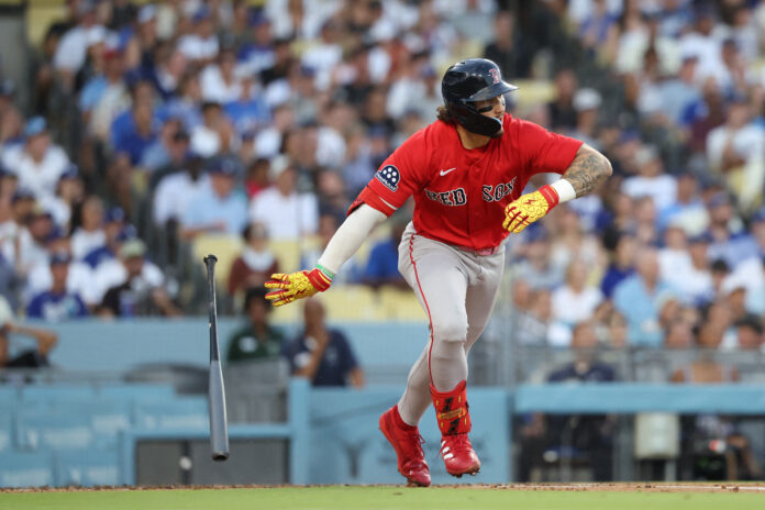 MLB Boston Red Sox at Los Angeles Dodgers Baseball player in a red Boston Red Sox uniform runs on the field, helmet on, with a bat on the ground and a blurred crowd in the stands.